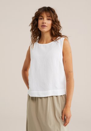 Žena s kučeravými hnedými vlasmi, oblečená v bielom tielku bez rukávov a béžovej sukni, stojaca pred jednoduchým svetlo béžovým pozadím.