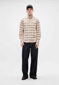 Camicia a quadri beige e marrone, abbinata a jeans in denim scuro a vita alta. Cappellino nero con testo bianco e scarpe nere.