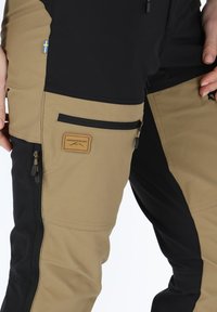 Bicolor broek in zwart en beige, met een ritszak, stoffen label en trekkoorden. Materiaal lijkt duurzaam en weersbestendig.