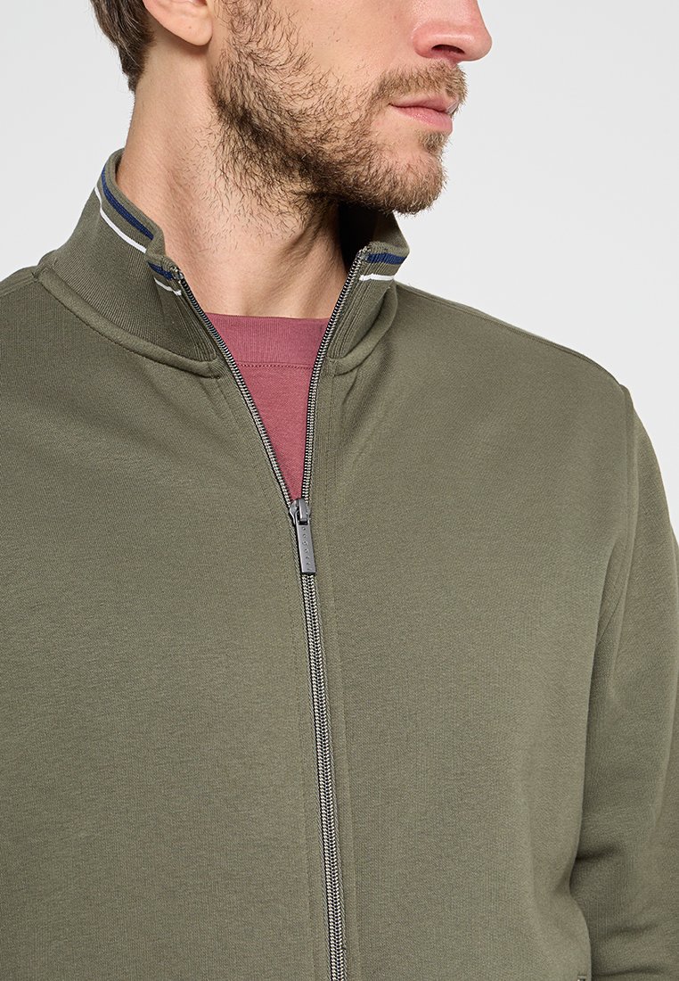 Uomo che indossa una giacca verde oliva con zip sopra una maglietta bordeaux, con la zip della giacca parzialmente chiusa e il colletto alzato.