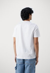 Camiseta blanca de algodón con mangas cortas, cuello redondo y una textura suave, presentada desde la parte trasera sobre un fondo liso.