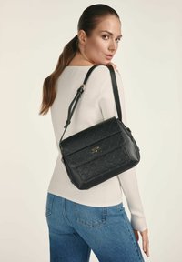 Kobieta z długimi brązowymi włosami związanymi w kitkę, ubrana w biały sweter w prążki i niebieskie dżinsy, trzymająca czarną pikowaną skórzaną torbę na ramię.