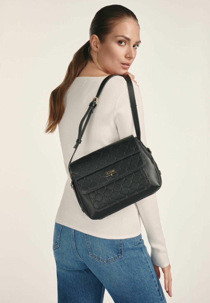 Kobieta z długimi brązowymi włosami związanymi w kitkę, ubrana w biały sweter w prążki i niebieskie dżinsy, trzymająca czarną pikowaną skórzaną torbę na ramię.