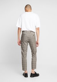 Man med beige rutiga byxor, vit oversized kortärmad skjorta och svarta finaskor, som står vänd bort från kameran mot en plain vit bakgrund.