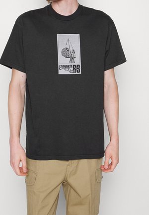 Mężczyzna w czarnej koszulce z grafiką dźwigu i głazu oraz beżowych spodniach typu cargo, stojący na białym tle.