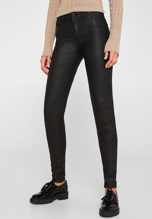 Kobieta ubrana w czarne obcisłe spodnie z powłoką, beżowy prążkowany sweter i czarne sznurowane buty stojąca na jasno szarej podłodze.