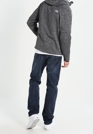 Mann trägt eine graue Kapuzenjacke, dunkelblaue Jeans und weiße Sneakers, steht mit dem Rücken zur Kamera auf einem einfachen weißen Hintergrund.