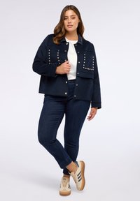 Jachetă bleumarin cu accente cu studs și buzunare frontale, asortată cu un top alb și blugi închisi, completată cu adidași de culoare deschisă.