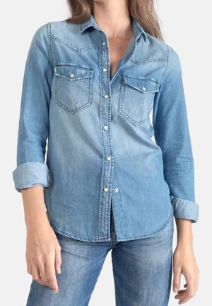 Lyseblå denimskjorte med perlemorsknapper, brystlommer og opkrogede ærmer, båret sammen med matchende blå jeans.