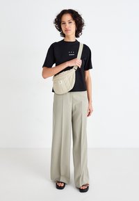 Beige gewatteerde crossbody tas met verstelbare schouderband, gecombineerd met een zwart T-shirt met korte mouwen en lichtgrijze wijde broek. Zwarte chunky sandalen.