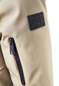 Beiger Stoffabschnitt eines Kleidungsstücks mit schwarzem "Polo Club Established 2012"-Aufnäher und schwarzem Reißverschluss mit der Aufschrift "Polo Club" an der Tasche.