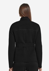 Svart bältejacka med krage, långa ärmar, mjuk textur och en skräddarsydd passform med synliga sömmar på baksidan.