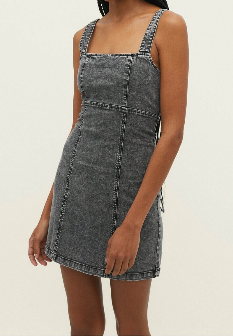 Femme portant une robe en denim gris cintrée sans manches avec un col carré et des détails de coutures visibles sur un fond clair.