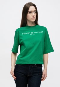 Πράσινο βαμβακερό t-shirt με κοντά μανίκια, με το λογότυπο "TOMMY HILFIGER EST. 1985" σε λευκό. Το τελείωμα τελειώνει πάνω από τη μέση.