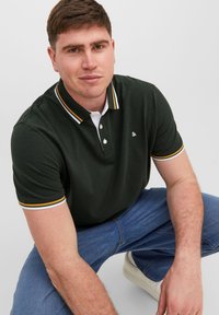Polo shirt verde scuro con colletto a righe bianche e gialle, patta con tre bottoni e piccolo logo sul petto. Indossato con jeans blu.
