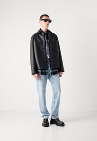Chaqueta negra de cuero con botones y dos bolsillos frontales, superpuesta a una camisa de cuadros, combinada con jeans de un azul claro y zapatos de plataforma negros.