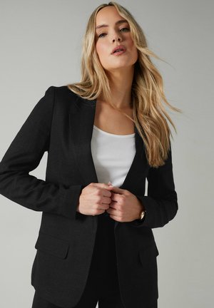 Black tailored blazer with a textured fabric, notched lapels, two front pockets, and a fitted silhouette, paired with a white top.