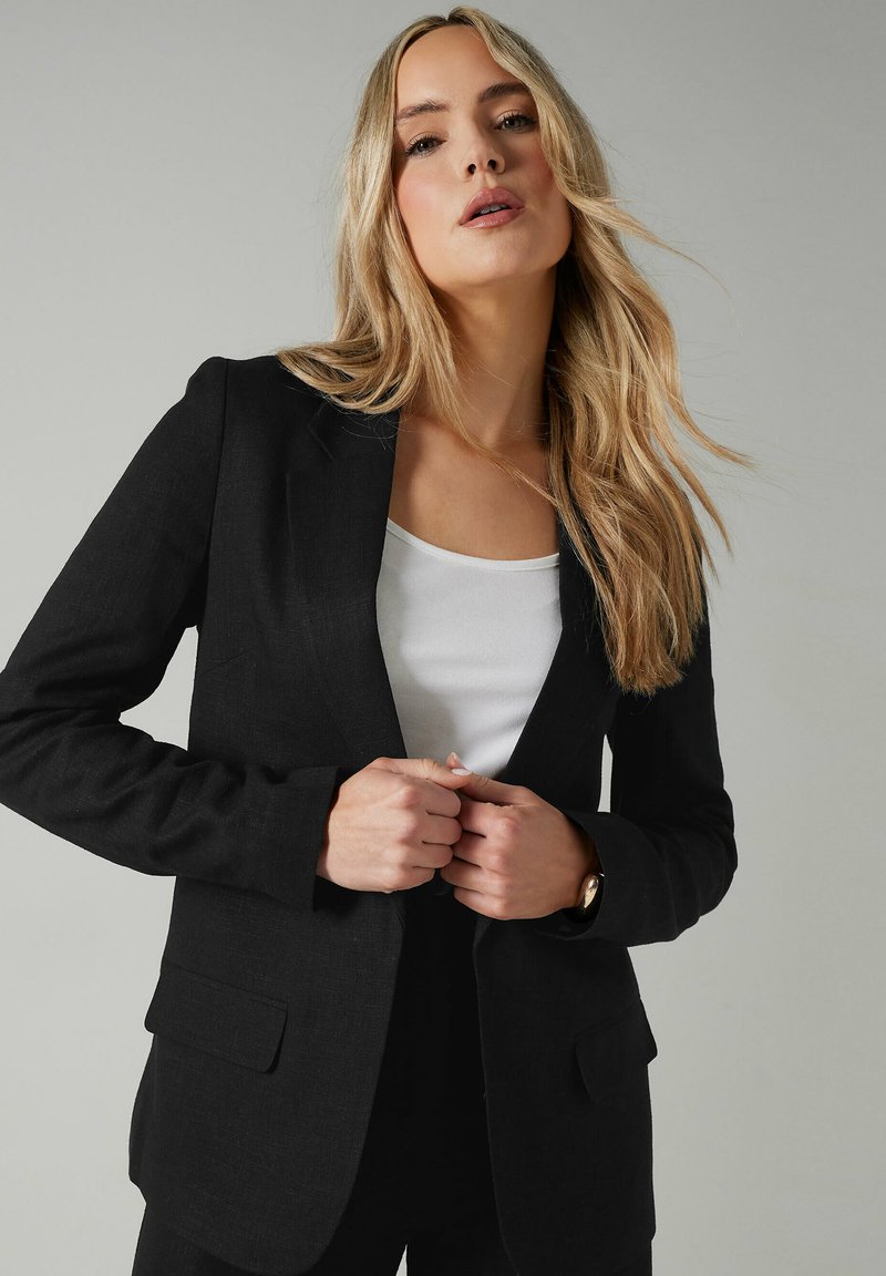Svart skräddarsydd kavaj i texturerat tyg, med slagkragar, två framfickor och en figursydd silhuett, kombinerad med en vit topp.
