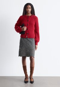 Roter, gerippter Strickcardigan mit schwarzen Knöpfen, kombiniert mit einem grauen Spitzenrock, braunen gerippten Socken und schwarzen spitzen Ballerinas.