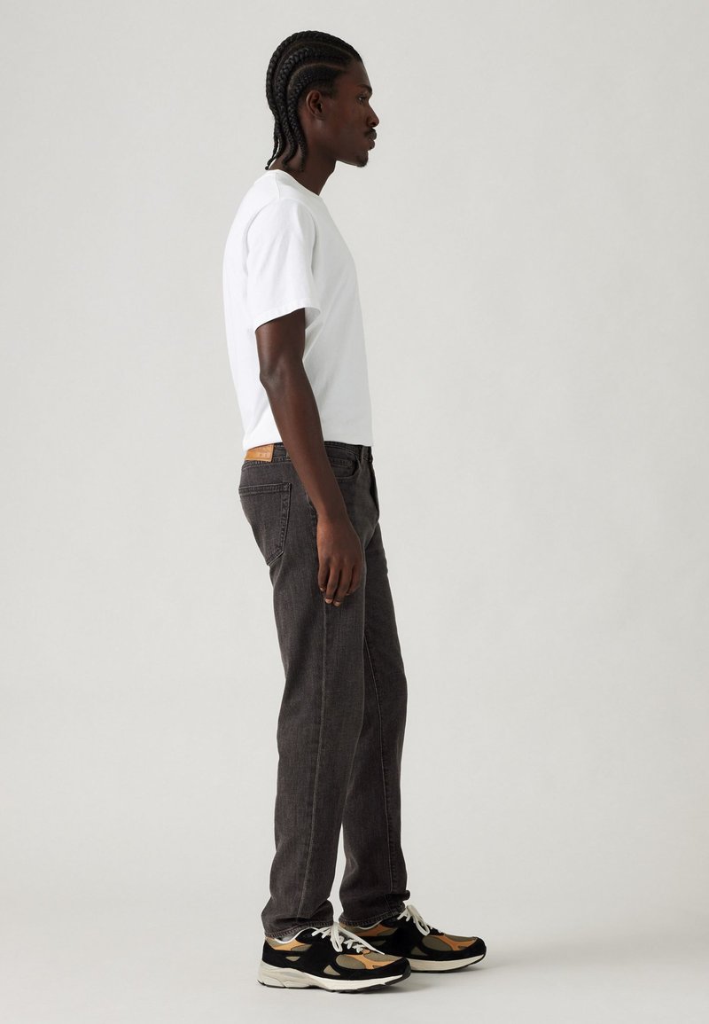 Camiseta blanca de manga corta, jeans de color gris oscuro y zapatillas negras multicolores. Peinado trenzado, de pie en perfil contra un fondo claro.