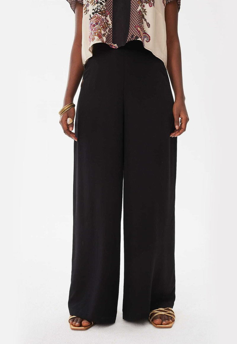 Zwarte wijde broek van gladde stof. Hoge taille, ruime pasvorm en volle lengte, gecombineerd met gouden accenten in de sandalen.