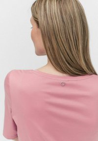 Kortärmad rosa T-shirt med en slät textur, rundad halslinning och en silverfärgad cirkulär accentdetalj på baksidan.