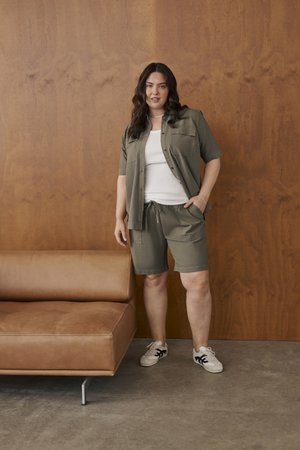 Femme aux longs cheveux foncés portant un short vert olive et une chemise par-dessus un top blanc, debout à côté d'un canapé en cuir beige contre un mur en bois.