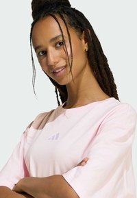 Svijetloružičasta majica s kratkim rukavima i okruglim izrezom. Ima suptilan dizajn ljubičastog logotipa na prsima. Mekana, glatka tekstura tkanine.