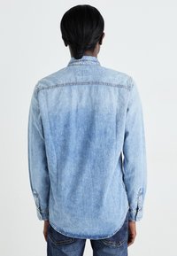 Camisa de denim azul claro con mangas largas, cuello puntiagudo y yugo en la parte posterior. Presenta detalles cosidos y un dobladillo ligeramente curvado.