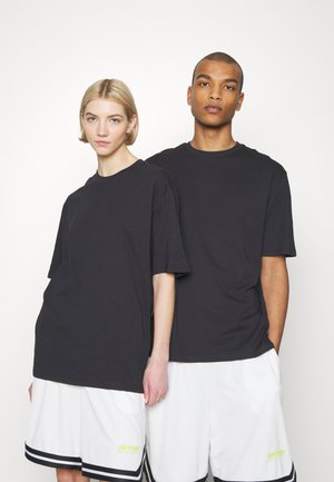 To modeller står side om side iført sorte, oversize T-shirts og hvide atletikshorts med sorte striber og gule logoer.