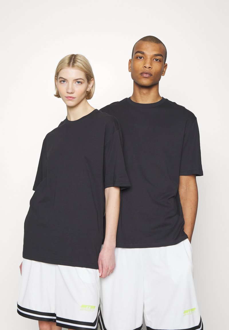 Dwie modelki stoją obok siebie, ubrane w czarne, oversize'owe koszulki oraz białe sportowe szorty z czarnymi paskami i żółtymi logo.