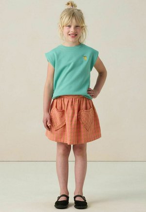 Lichtblauw T-shirt met korte mouwen en een klein geel ontwerp, gecombineerd met een roze geruite rok met hartvormige zakken. Zwarte schoenen.