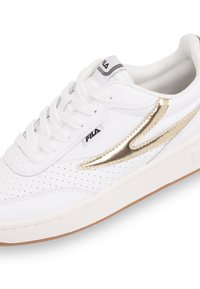 Sneaker bianca con dettagli metallici dorati, forature decorative e suola in gomma texturizzata. Lacci e logo FILA in nero.