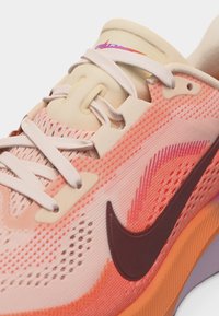Gros plan sur une chaussure de course rose et beige avec des lacets blancs, une tige en mesh, une semelle orange, et un logo Nike swoosh brun foncé sur le côté.