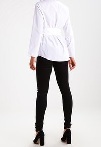 White long-sleeve shirt with a tie waist, paired with fitted black trousers and black high heels. Smooth fabric with a clean design.