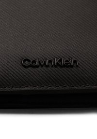 Cartera de cuero texturizado negro con un logo "Calvin Klein" en relieve. El exterior presenta suaves líneas horizontales y un acabado liso.