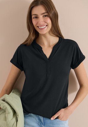 Femme souriante aux longs cheveux bruns portant un t-shirt noir à manches courtes et un jean bleu clair, tenant une veste vert clair.