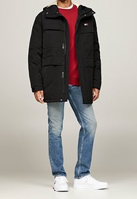 Czarna pikowana kurtka parka z odpinanym kapturem, przednimi kieszeniami i beżową podszewką, noszona na czerwonej bluzie i niebieskich dżinsach. Białe trampki.