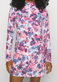 Kvinna iklädd en långärmad tröja med kvart-zip och matchande kjol med ett abstrakt blomtryck i rosa, blått och lila.