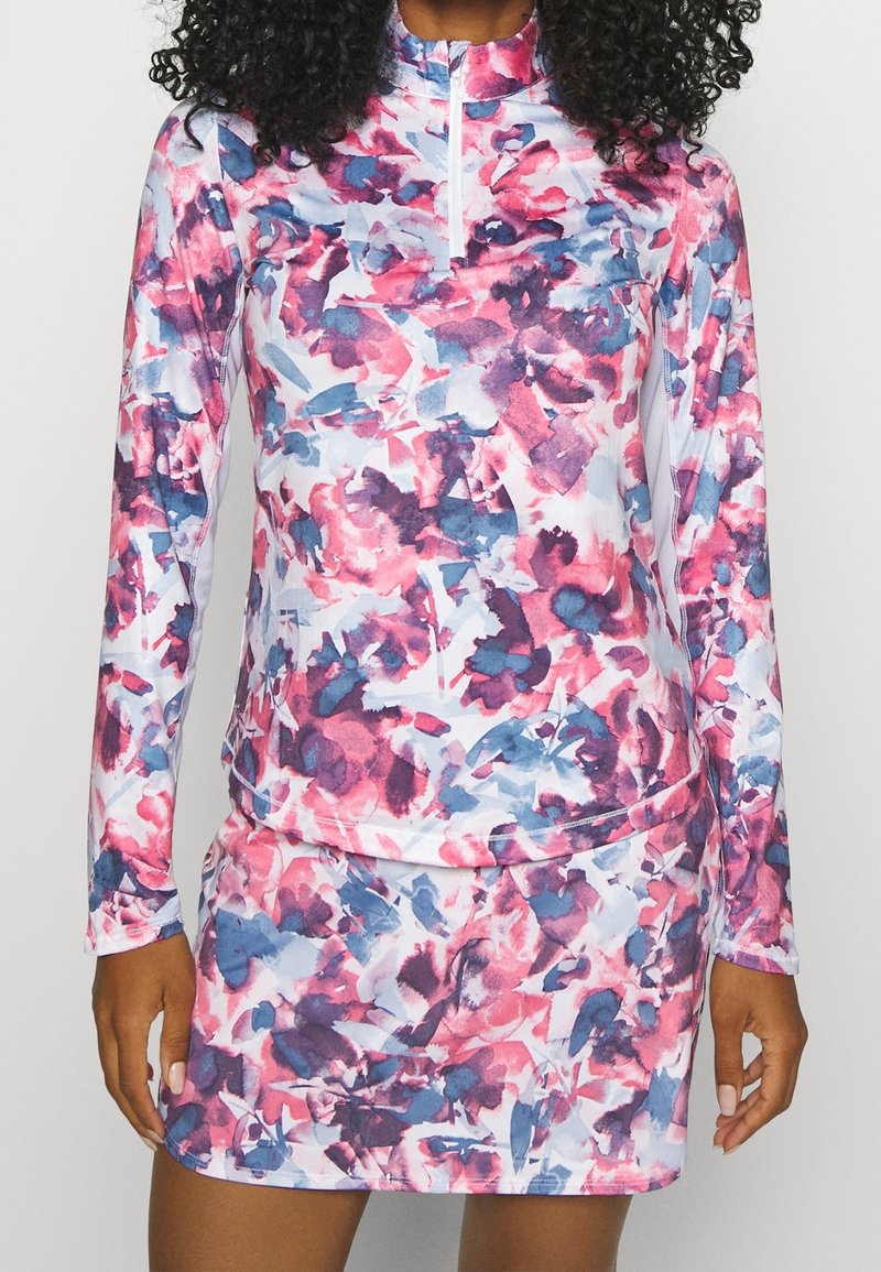 Kvinna iklädd en långärmad tröja med kvart-zip och matchande kjol med ett abstrakt blomtryck i rosa, blått och lila.