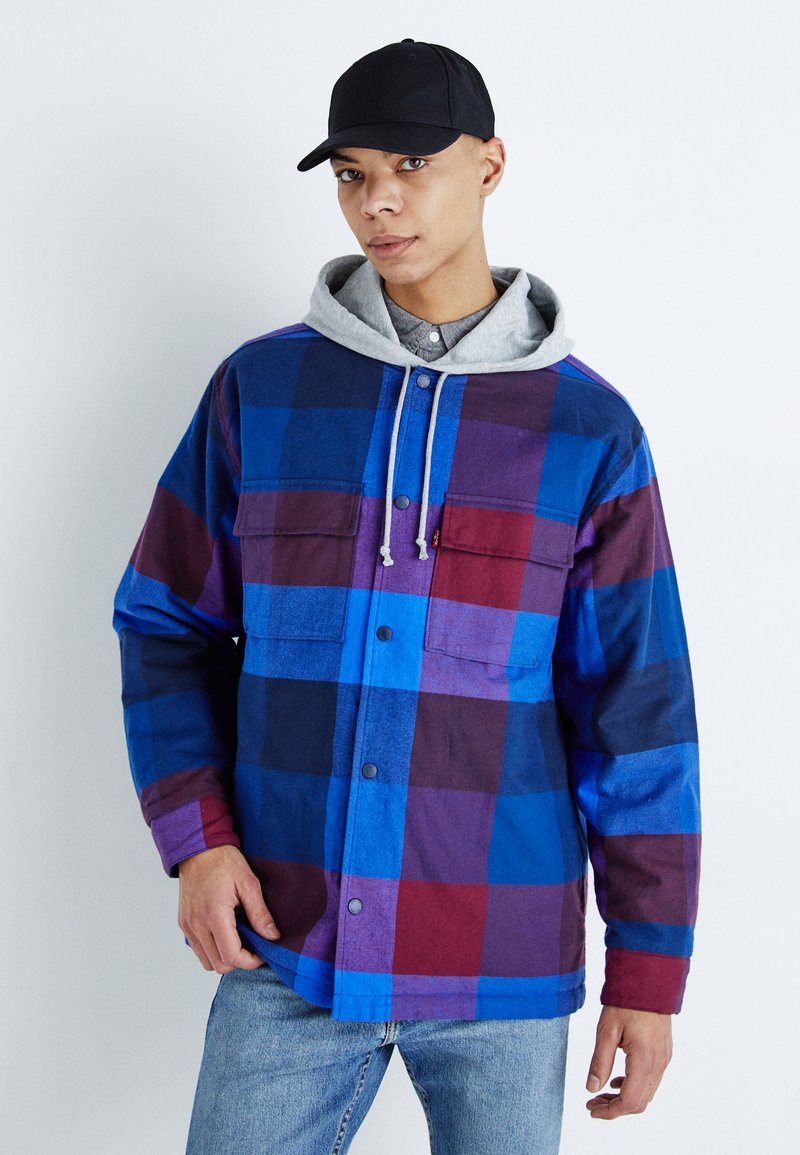 Kariertes Flanelljackett in Blau, Lila und Burgunderrot. Verfügt über zwei Brusttaschen, Knopfverschlüsse und einen grauen Kapuzenpullover darunter.