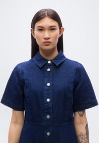 Φόρεμα πουκάμισο από τζιν σε σκούρο μπλε με γιακά, κοντά μανίκια και ασημένια κουμπιά κατά μήκος του μπροστινού μέρους. Διαθέτει εφαρμοστή σχεδίαση.