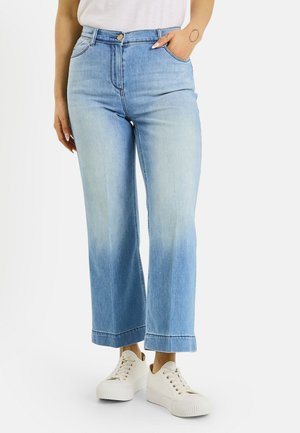 Mujer con vaqueros cortos de pierna ancha de color azul claro con dobladillos enrollados, zapatillas blancas y una camiseta blanca, mano en el bolsillo, de pie sobre fondo blanco.