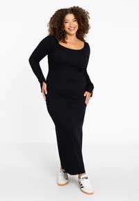 Langes schwarzes Kleid mit rundem Ausschnitt und langen Ärmeln, das eine taillierte Silhouette aufweist. Gestylt mit weißen Sneakers. Weicher, elastischer Stoff.