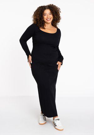 Langes schwarzes Kleid mit rundem Ausschnitt und langen Ärmeln, das eine taillierte Silhouette aufweist. Gestylt mit weißen Sneakers. Weicher, elastischer Stoff.