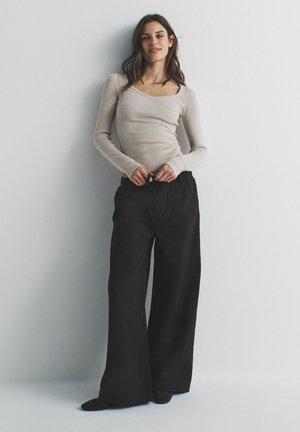 Frau mit langen braunen Haaren steht vor einer weißen Wand, trägt ein beige geripptes Langarmoberteil und schwarze Hosen mit weitem Bein, Hände in der Taille.