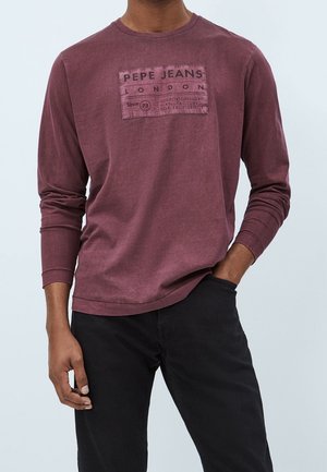 Homme portant une chemise manches longues bordeaux avec le texte "PEPE JEANS LONDON" et un pantalon noir, main dans la poche, devant un fond uni.