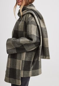 Modell som bär en rutig kappa i nyanser av grått och beige, med en matchande scarf hängande över axeln, mot en neutral bakgrund.