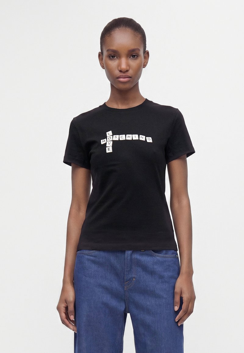 Γυναίκα που φοράει μαύρο t-shirt με σχέδιο σταυρόλεξου που σχηματίζει το "Love Moschino" και μπλε τζιν, στέκεται μπροστά σε λευκό φόντο.