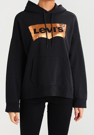 Personne portant un sweat à capuche noir avec une poche avant et un logo Levi's cuivre brillant sur la poitrine.
